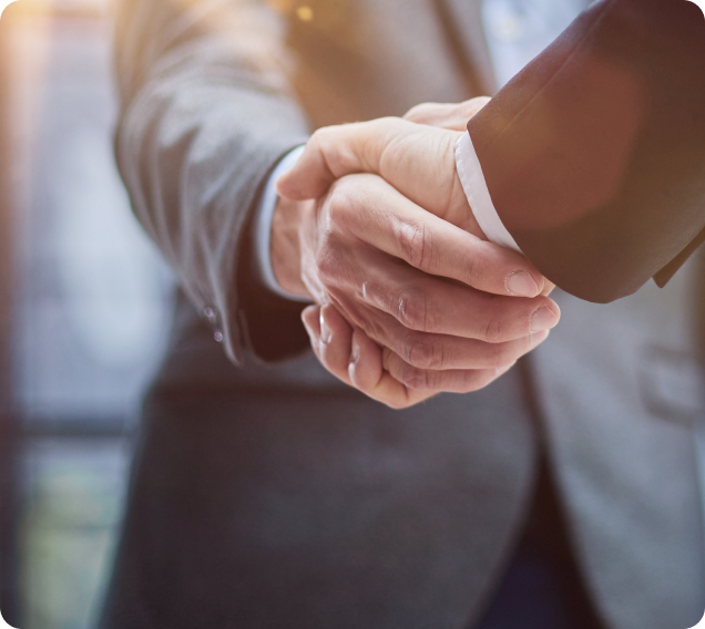 This image shows a cropped scene of two hands set in a handshake grip. The left hand belongs to a man wearing a grey suit and the right hand which is set in the fore ground of the image belongs to a man in a black suit.