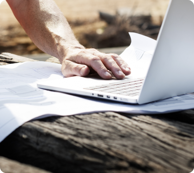 The image shows a cropped scheme of one hand holding down documents on a wooden table, there is a laptop off to the right but mostly out of view. Both the papers and the laptop are white. The image is taken outside with the background blurred by yellows and browns.