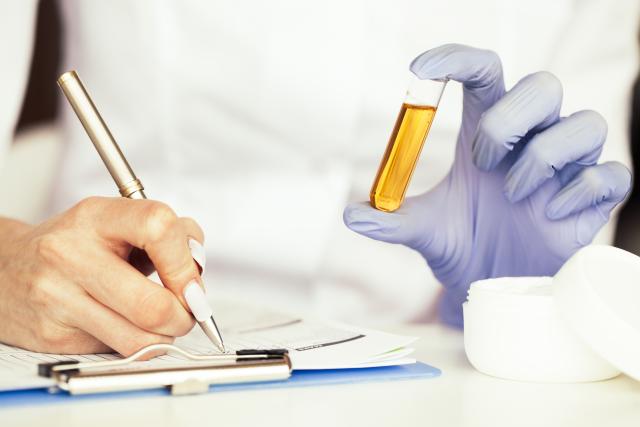 The image shows a person wearing gloves, holding a test tube with a yellowish liquid which is being tested. The person is also holding a pen and appears to be writing or marking on a piece of paper or a clipboard data about the liquid testing. This scene suggests a laboratory setting where testing data of substances is being conducted. The person is likely a scientist or technician involved in the analysis and documentation of the results.