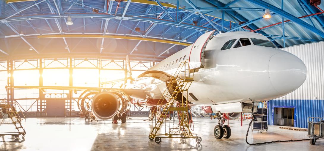 This image shows and aircraft in an aircraft hangar. The aircraft is off centre to the right of the image, with its wing cutting across the remainder of the frame from right to left. The door one passenger door and cargo hold door of the aircraft are open with yellow workers steps leading up to them. The aircraft hangar has a blue industrial roof and a large window running behind the aircraft with light coming through, the floor is concrete with a clean shine to it.