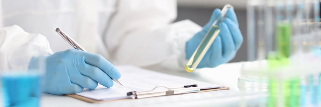 The image shows a pair of hands in blue plastic gloves in the left hand is a test tube with light yellow liquid, in the right is a silver pen. The pen tip is resting on a clip folder as if to write. The focus of the image is on the pen and the test tube with the background of the persons arm and white laboratory coat is blurred.