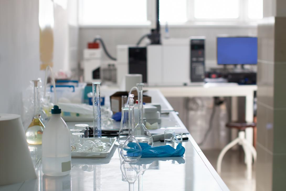 The image depicts a chemistry laboratory with a focus on a table laden with various glass bottles and laboratory equipment. In the background, there are additional pieces of equipment, including what looks like a centrifuge and a computer monitor, which further emphasises the scientific nature of the setting.