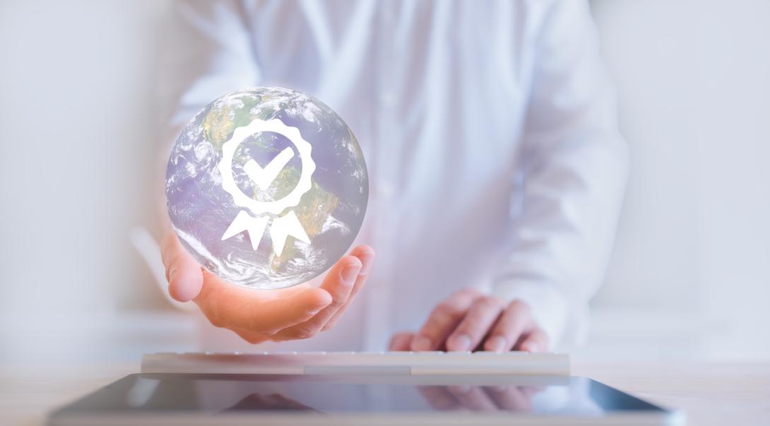 The image depicts a pair of hands gently holding a transparent globe with a detailed representation of the Earth. The globe is adorned with a check symbol that signifies quality assurance, guarantee, standards, certification, and standardisation. The background features a blurred figure of a person wearing a white lab coat, suggesting a professional or scientific setting.